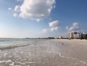 A beach with a clear sky and a few clouds. There are buildings in the background.