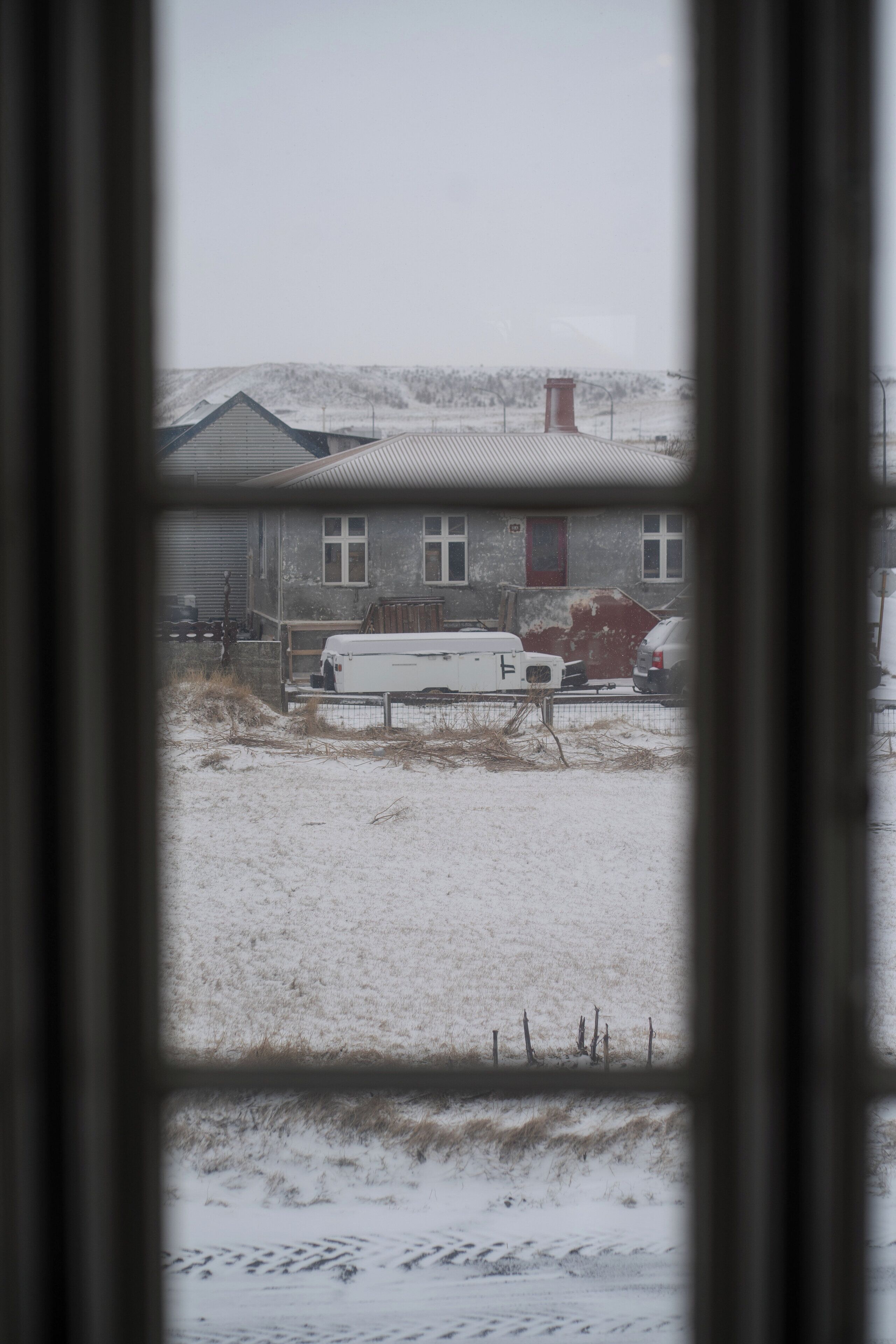 Vista a través de una ventana que muestra una casa con patio nevado, una camioneta blanca estacionada y un coche rojo.