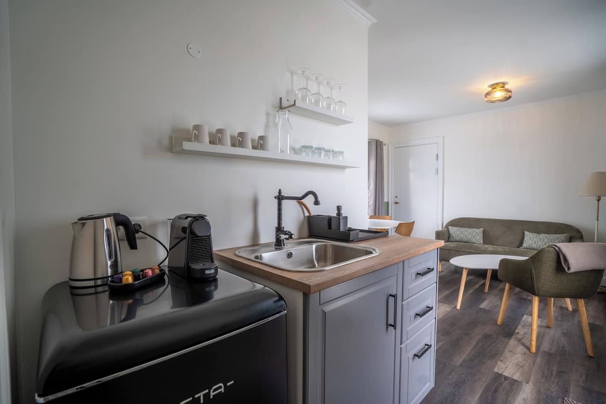 Cocina compacta con cafetera, hervidor y tostadora. Hay repisas sobre el lavadero con cristalería. Se ve un pequeño comedor con una mesa y sillas al fondo.