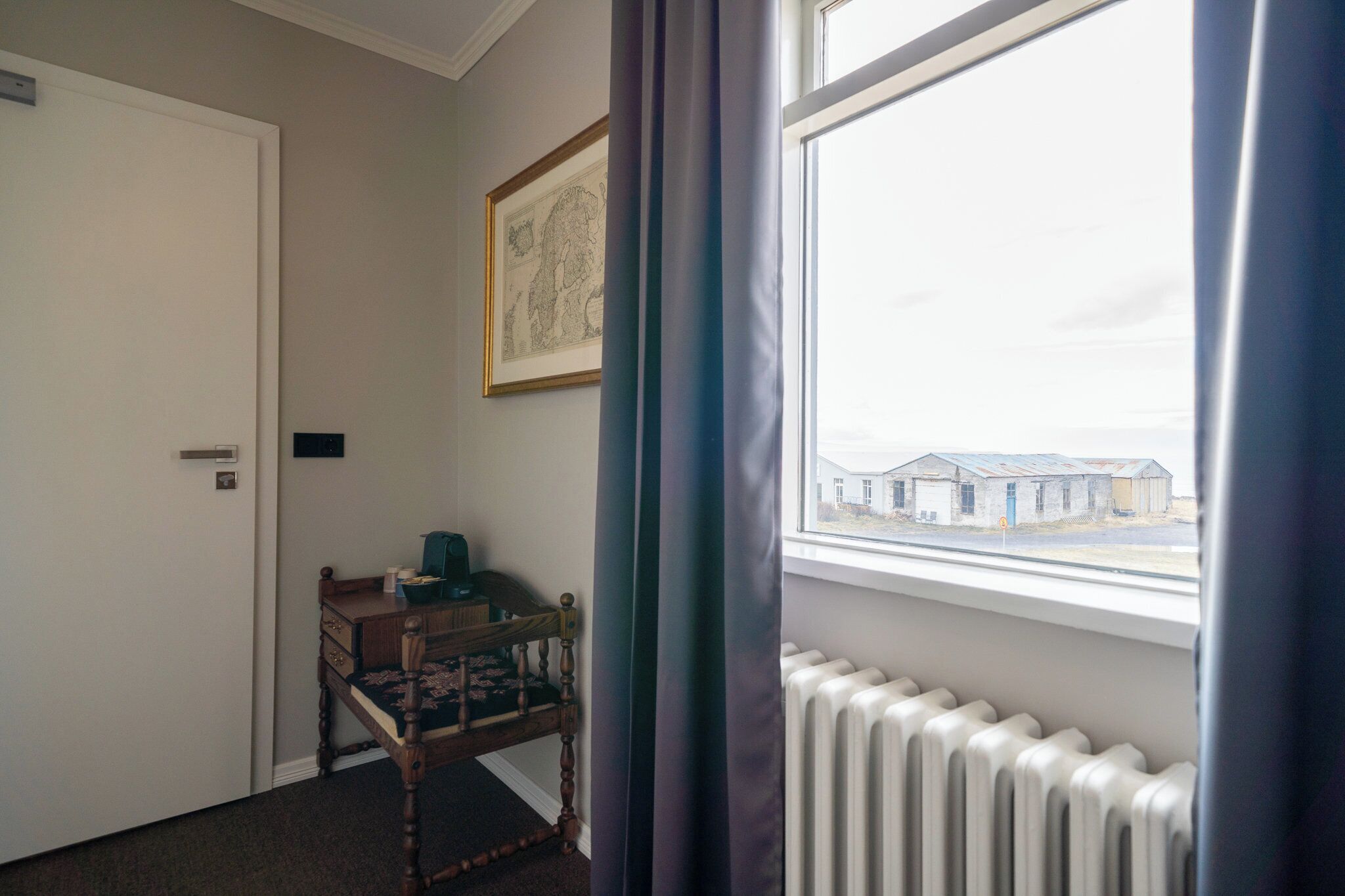 A room with a bed, a small table, a radiator, a window with a view of buildings, and a door.