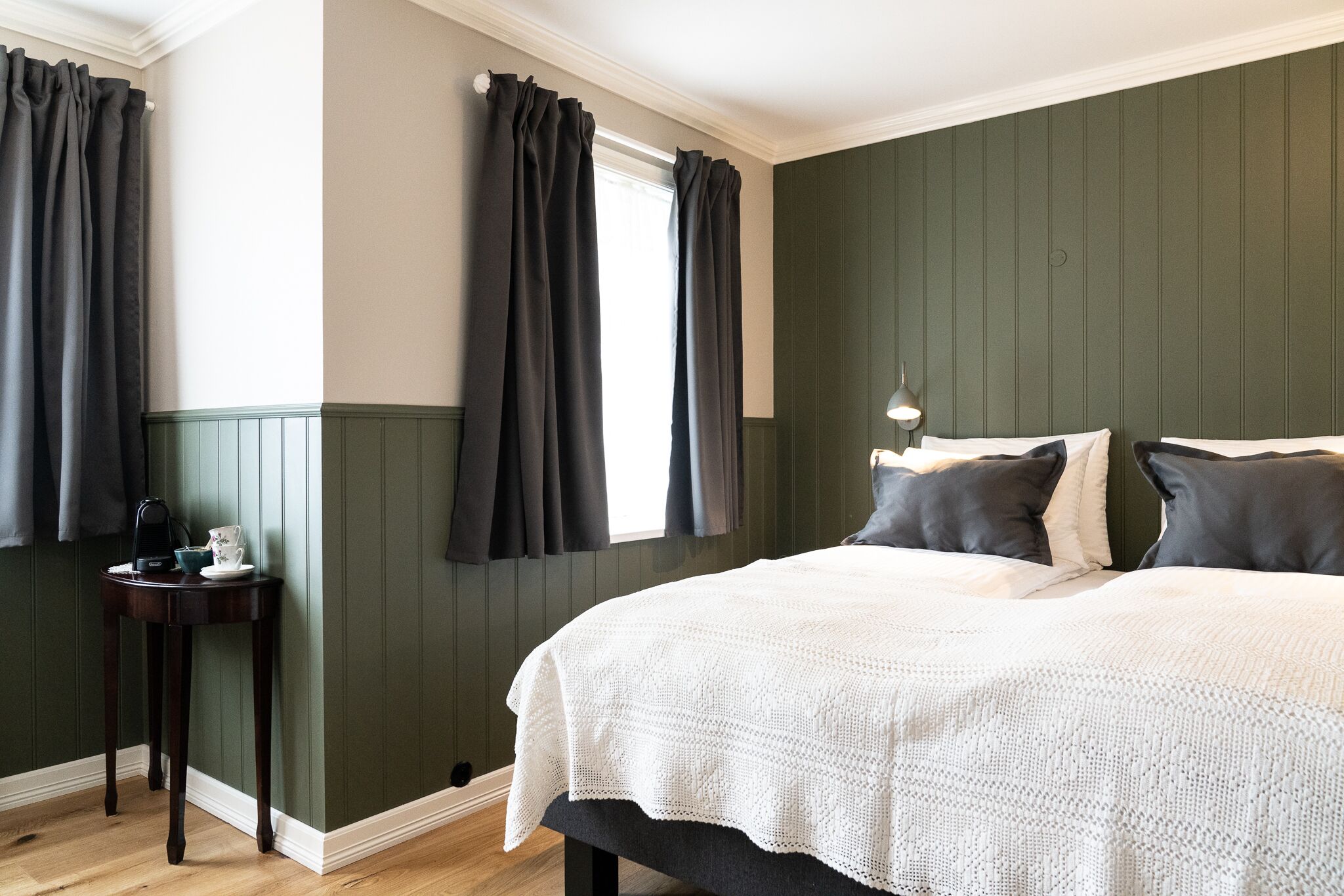 Un dormitorio con una cama grande, cortinas oscuras, una mesita de noche con lámpara y paredes de paneles verdes.