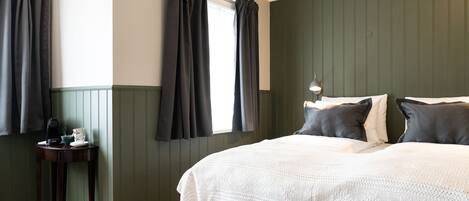 Un dormitorio con una cama grande, cortinas oscuras, una mesita de noche con lámpara y paredes de paneles verdes.