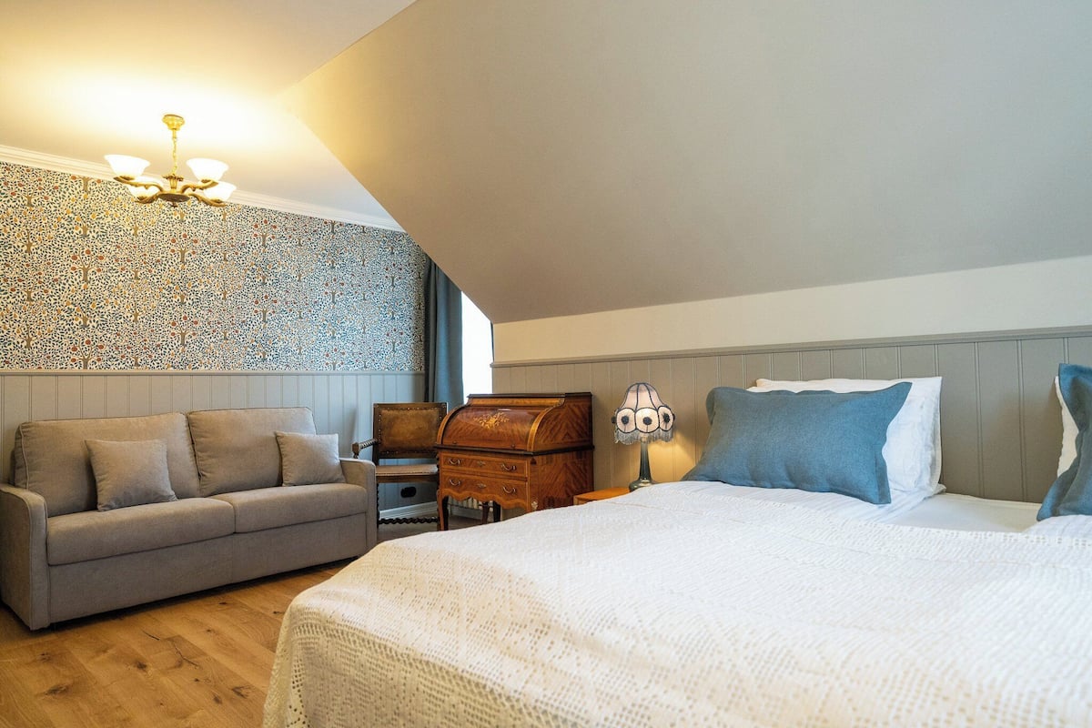 Un dormitorio con cama, sofá, un cofre de madera y una lámpara.