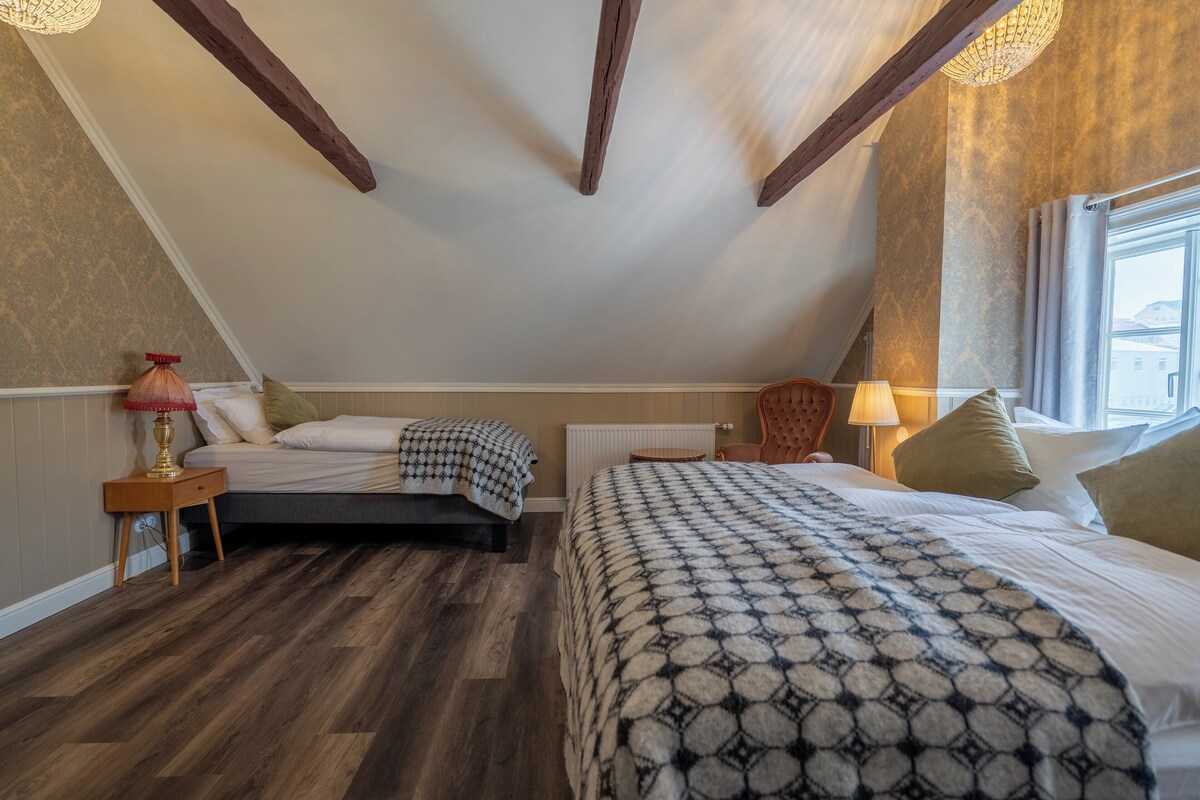 A bedroom with two beds, wooden beams on the ceiling, a radiator, and a window with curtains.