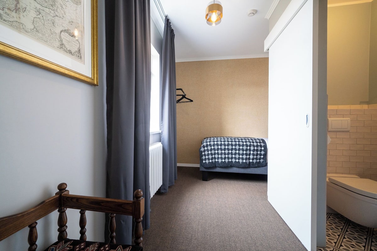 Un pasillo estrecho con una silla de madera, un radiador, una cama con manta estampada y un perchero.