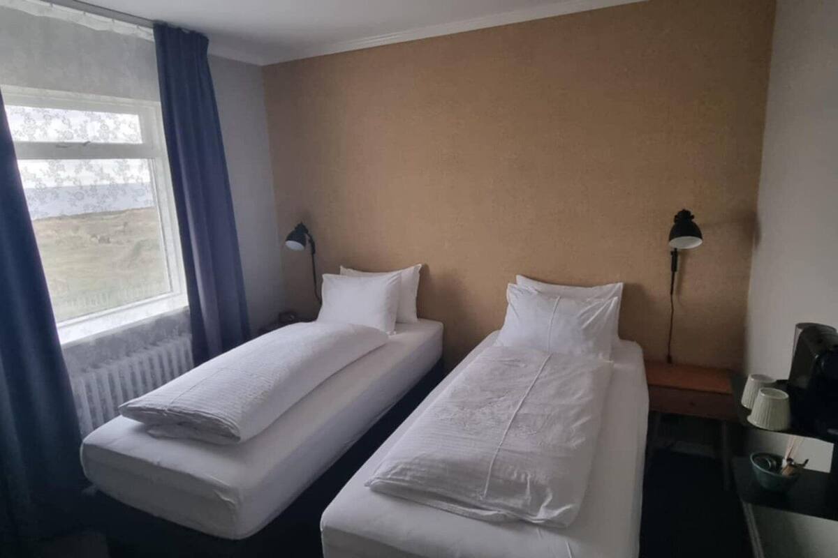 Una habitación de hotel con dos camas individuales, una mesita de noche con lámpara y una ventana con cortinas.