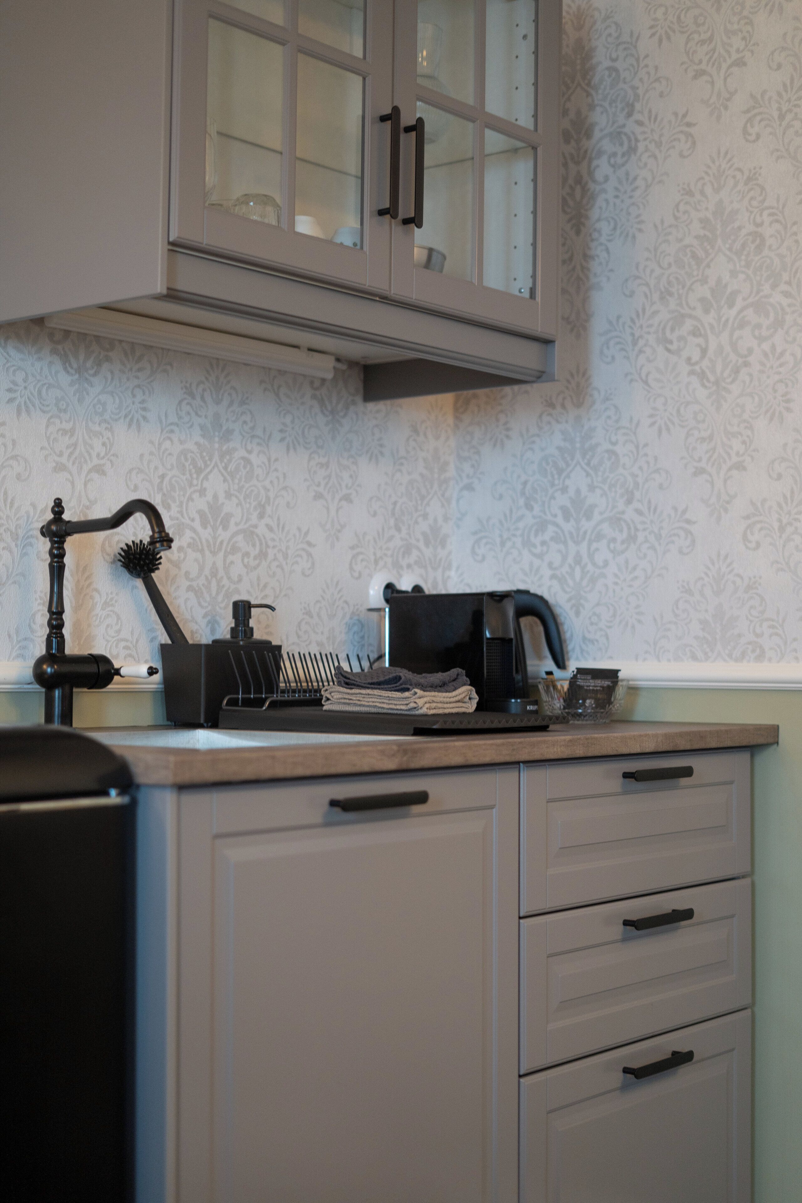 Eine Küche mit Kaffeemaschine, Wasserkocher und Spüle. Über der Arbeitsplatte befinden sich Schränke und die Wand ist mit einem gemusterten Wandpapier versehen.