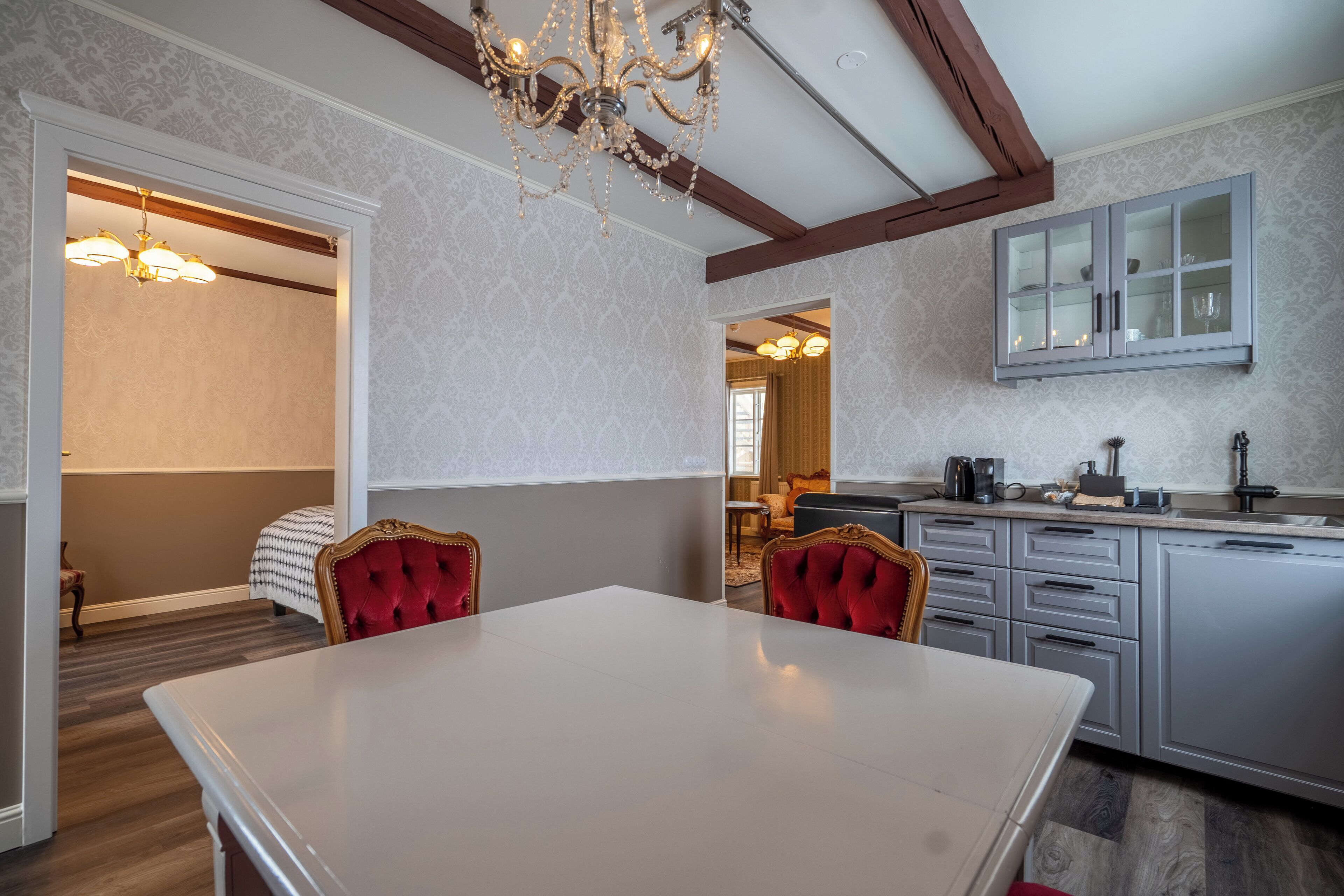 A dining area with a white table and red upholstered chairs, a chandelier, and a kitchen with grey cabinets and a built-in microwave.