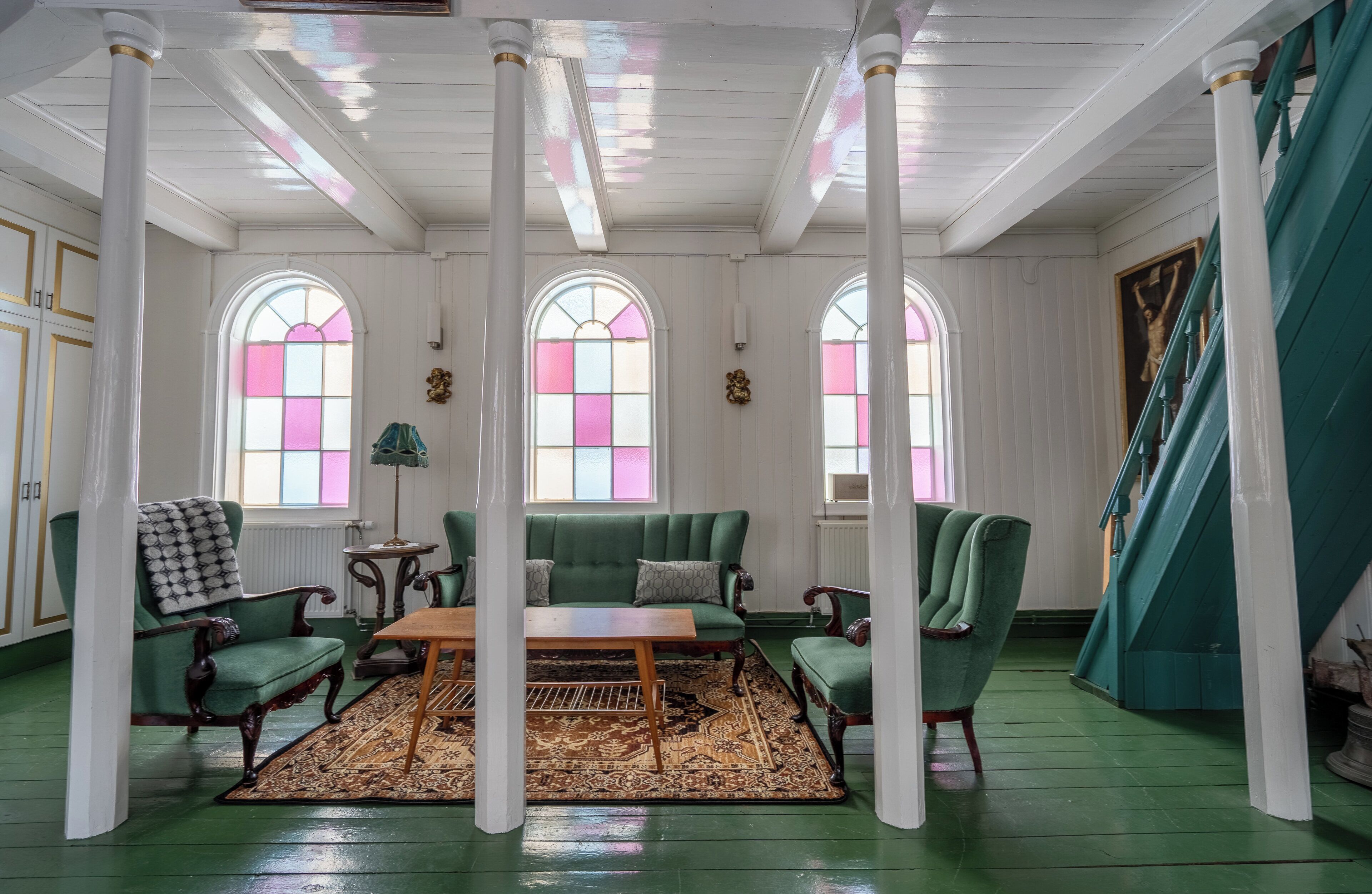 A room with a green sofa set, a wooden coffee table, and a patterned rug. There are two arched windows with pink and white panes.
