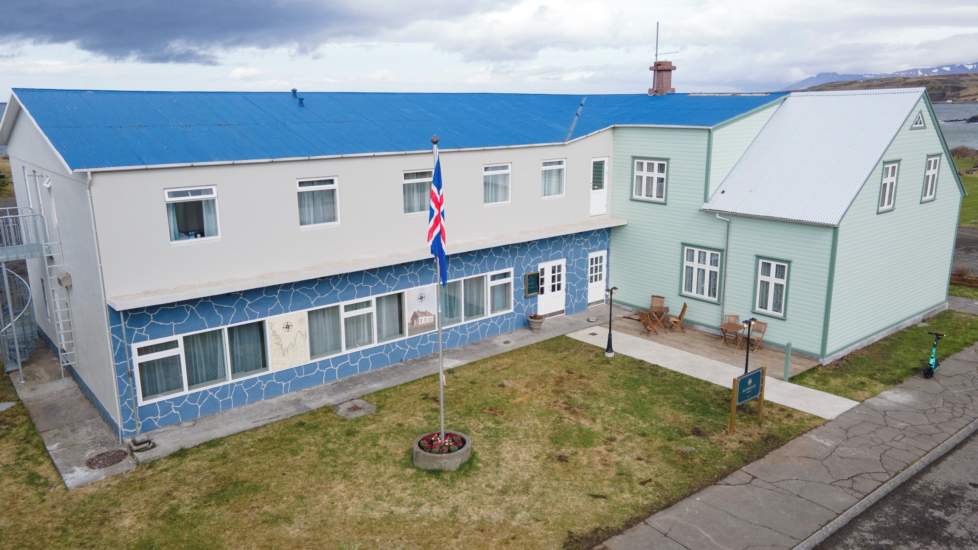 Un edificio de dos pisos con techo azul, un asta con bandera y un pequeño jardín.