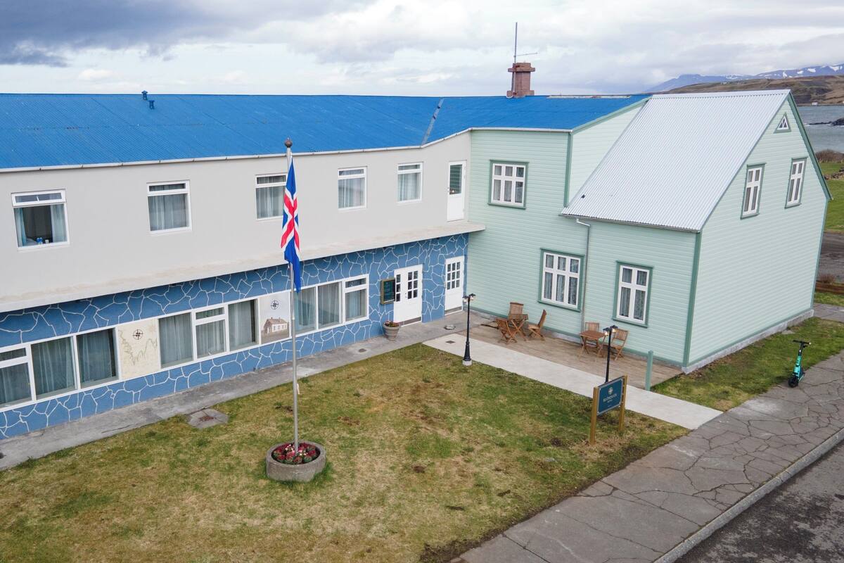 Un edificio de dos pisos con techo azul, un asta con bandera y un pequeño jardín.