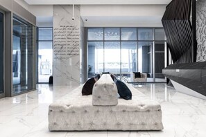 A modern lounge area with a plush sofa, a marble pillar with text, and large windows offering a view. - Boho-Chic Apt w Canal view (Dubai)