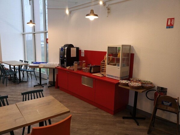 Un café moderno con mostrador rojo, máquinas de café y un refrigerador. Hay mesas y sillas de madera, y ventanas amplias que ofrecen luz natural.