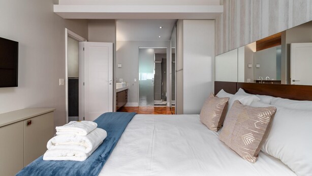 Habitación de hotel moderna con una cama grande, una mesita de noche y un espejo encima. Se ve un baño a través de una puerta abierta al fondo.