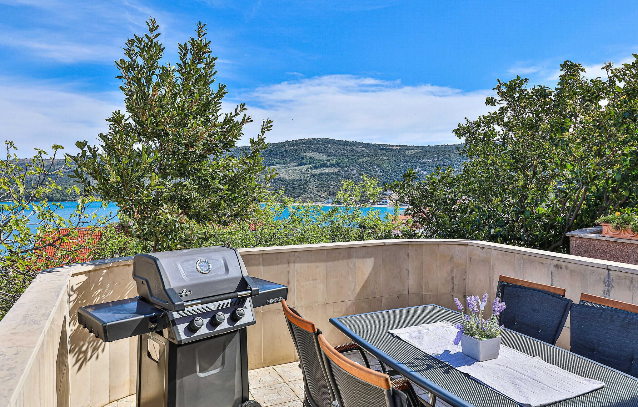 Patio esterno con barbecue, tavolo e sedie, affacciato su un panorama che include un lago e delle colline.