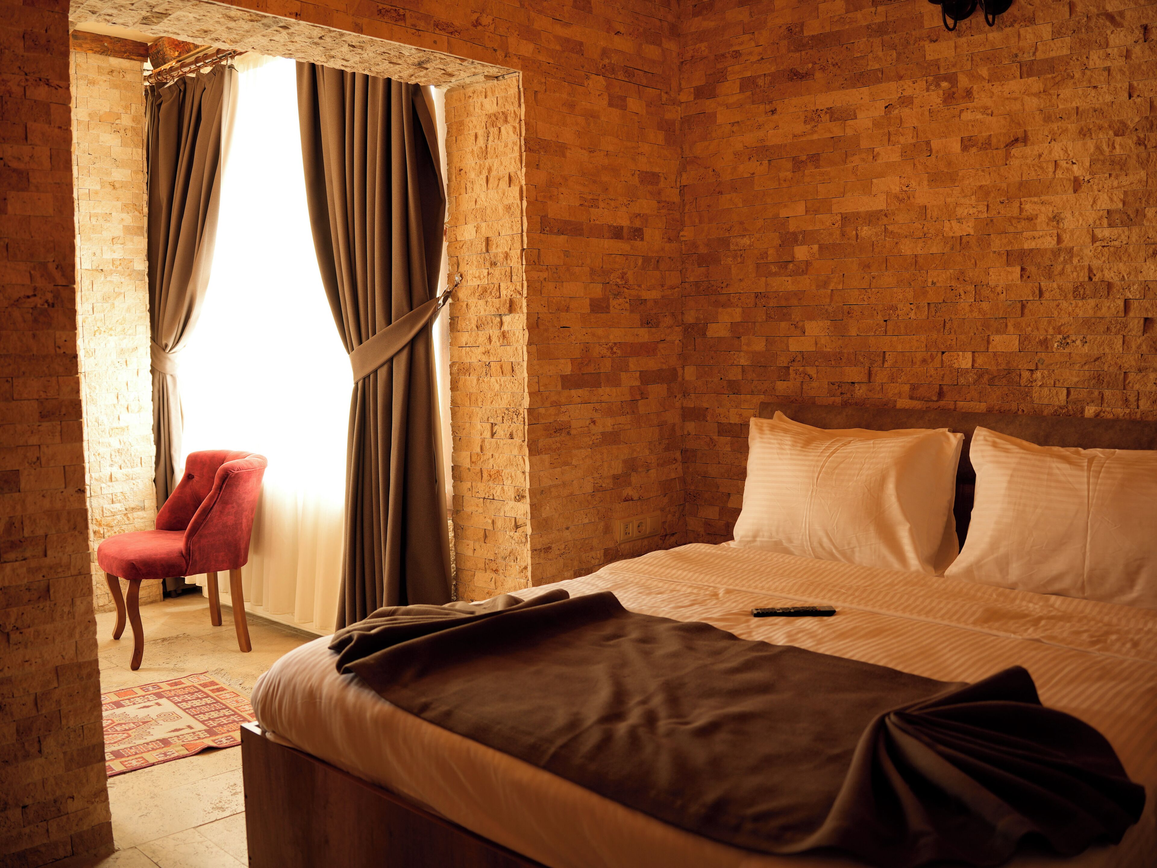 Una habitación con pared de ladrillo, una cama con almohadas blancas, una silla roja y una ventana con cortinas.