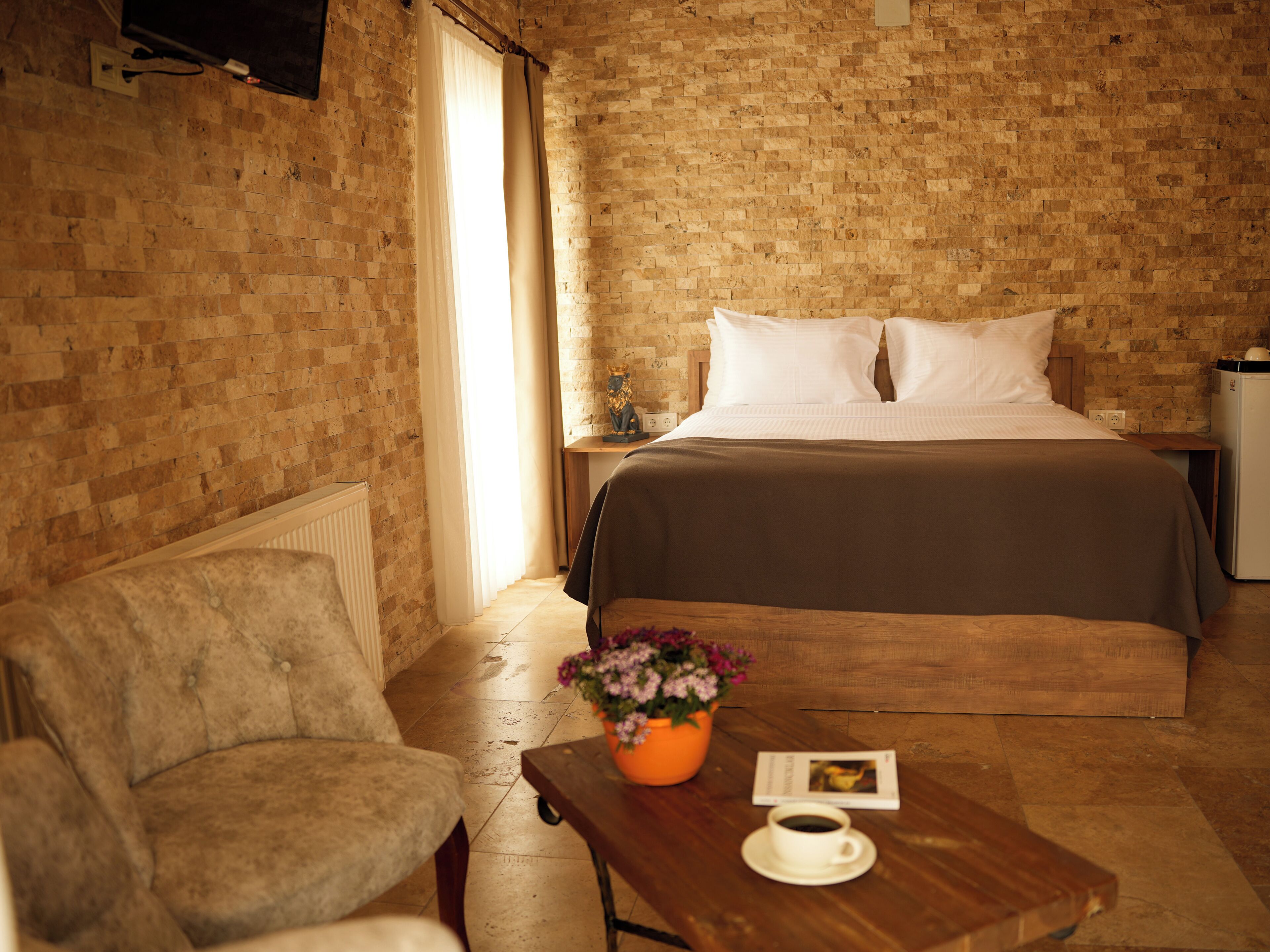 A hotel room with a brick wall, a bed with white linens, a wooden chair, a small table with a cup of coffee, and a potted plant.