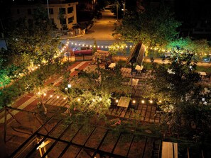 Un área de estar al aire libre con mesas y sillas, rodeada de follaje e iluminada con guirnaldas.
