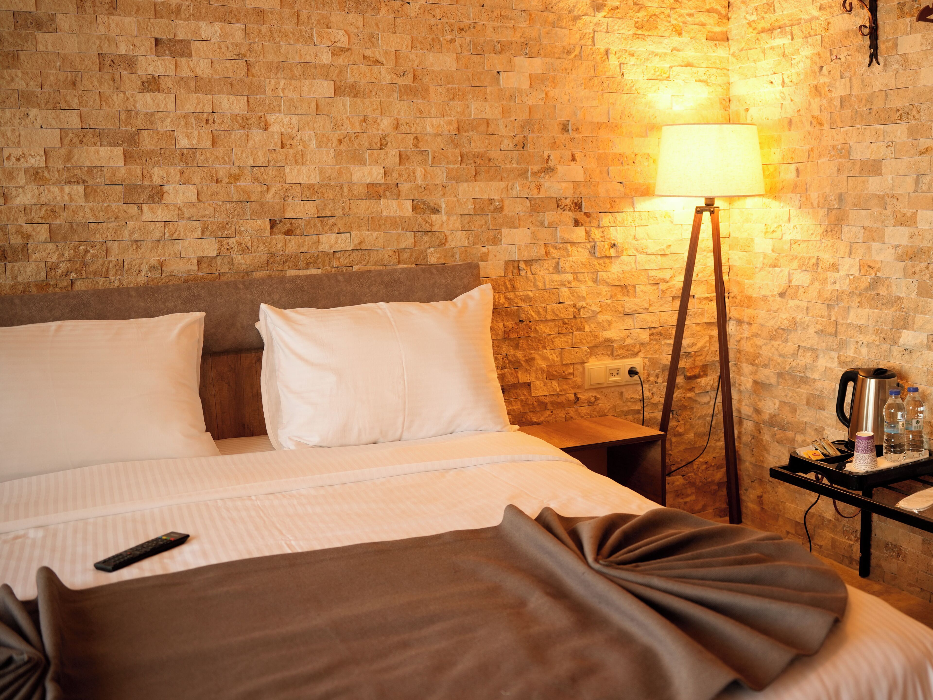 A hotel room with a brick wall, a bed with white linens, a bedside table with a lamp, and a nightstand with a kettle and bottles.