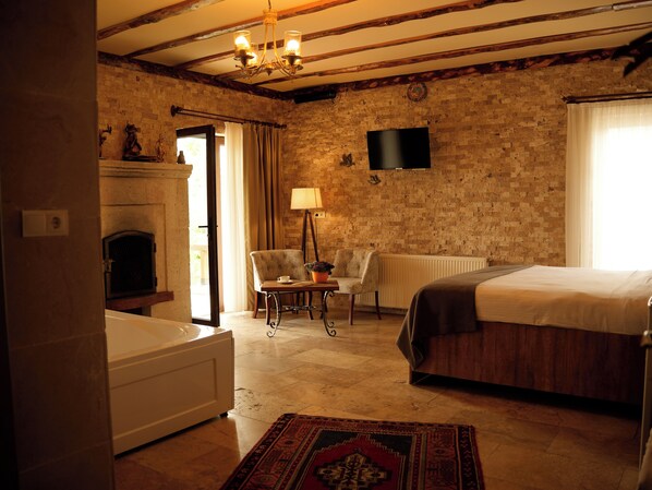 A hotel room with a brick wall, a fireplace, a bed, a chair, a table with a lamp, and a TV mounted on the wall.