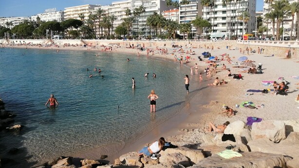 Een strand met helder water, mensen die zwemmen en zonnebaden, en gebouwen op de achtergrond.