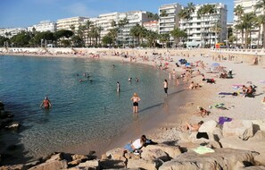 En strand med klart vatten, där folk badar och solar, och med byggnader i bakgrunden.