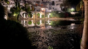 Vista nocturna de un edificio de estilo mediterráneo tradicional con varios balcones, un estanque con nenúfares y vegetación circundante.