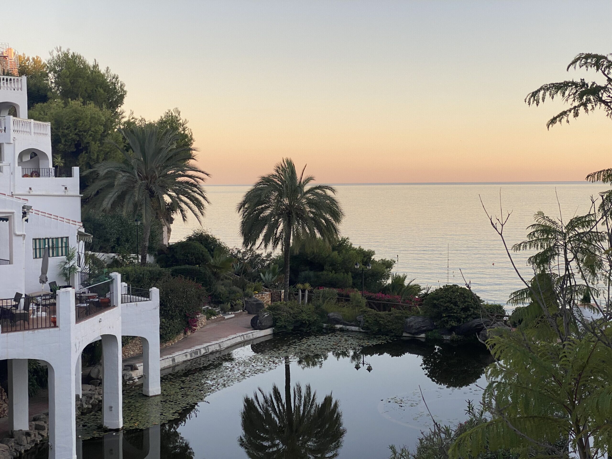 Une propriété paisible située au bord de l’eau, avec un bâtiment blanc doté de balcons en arc de cercle, un étang parsemé de nénuphars et une vue sur la mer au coucher du soleil.