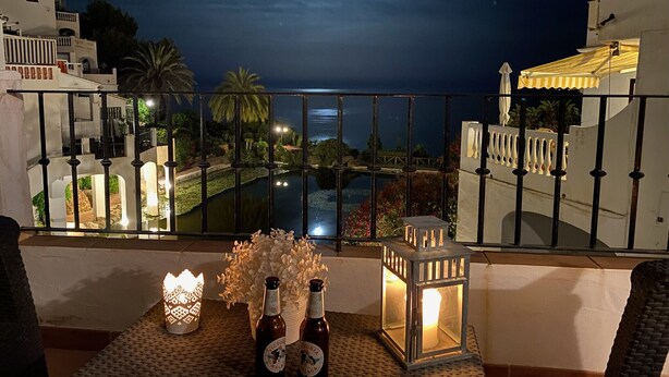 Un balcón con vistas a una piscina y al cielo nocturno. Hay dos botellas y un farol en la mesa.