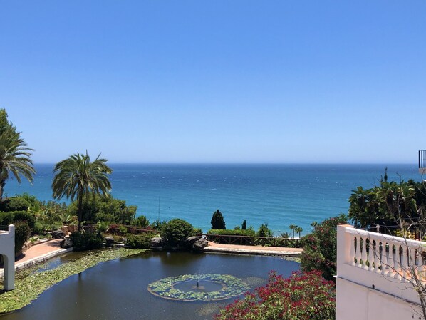A view of a calm body of water with lily pads and a fountain, surrounded by lush greenery and palm trees, leading to a clear blue sea. - AP231 - Capistrano Playa (Nerja)