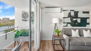 Un soggiorno moderno con un divano grigio, una lampada da terra e un grande quadro astratto. C'è un balcone con vista su edifici e vegetazione.