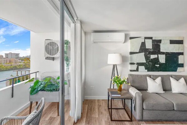 Un salón moderno con un sofá gris, una lámpara de pie y un cuadro abstracto grande. Hay un balcón con vistas a edificios y zonas verdes.
