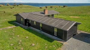 En modern, enplansbyggnad med ett sluttande tak, stora fönster och en skorsten. Den ligger på en gräsklädd kulle med utsikt över havet i bakgrunden.