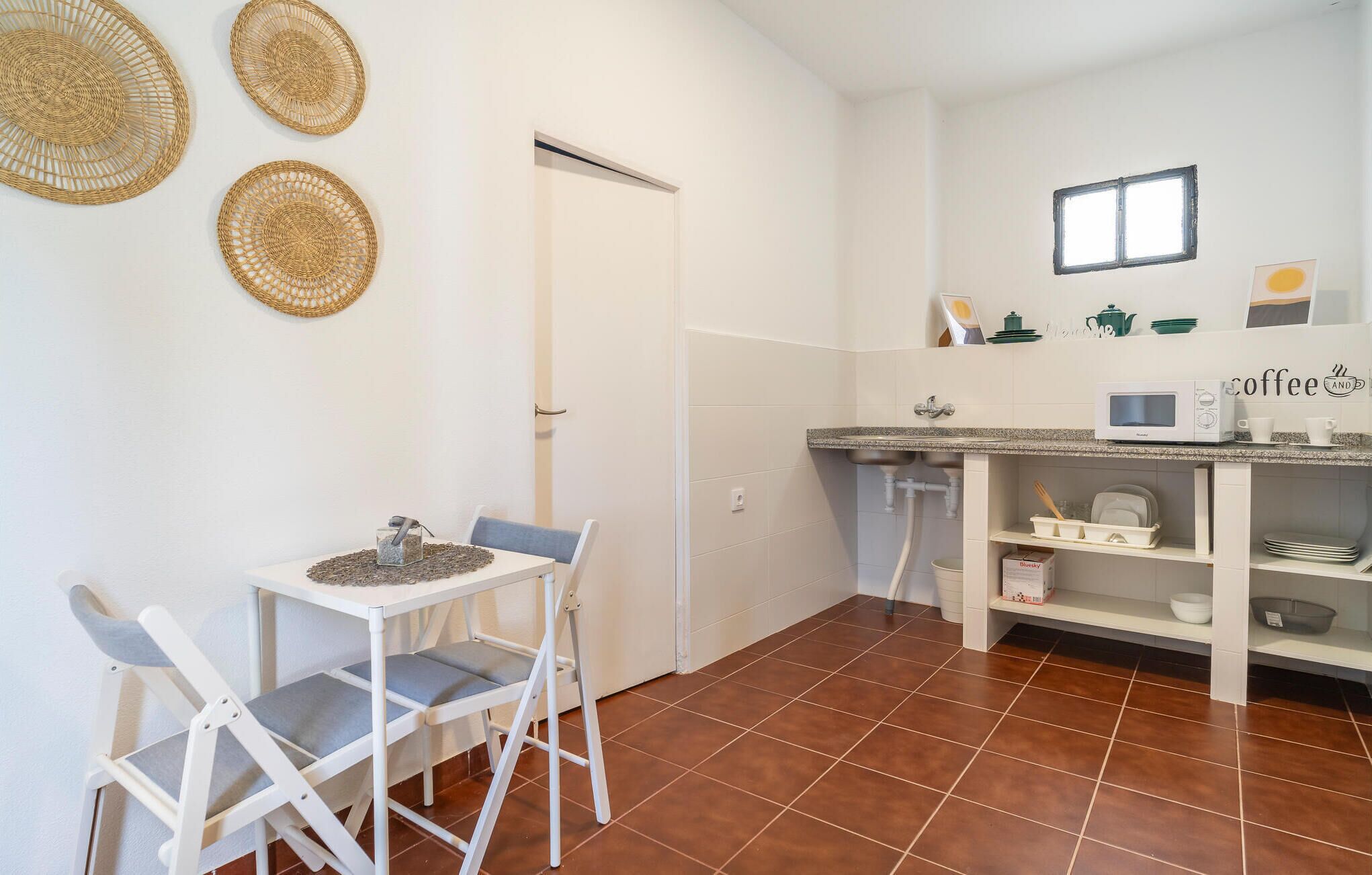 Een compacte keuken met witte kastjes, een magnetron en een koffiehoekje. Drie geweven placemats hangen aan de muur en er staat een klein tafeltje met twee stoelen.