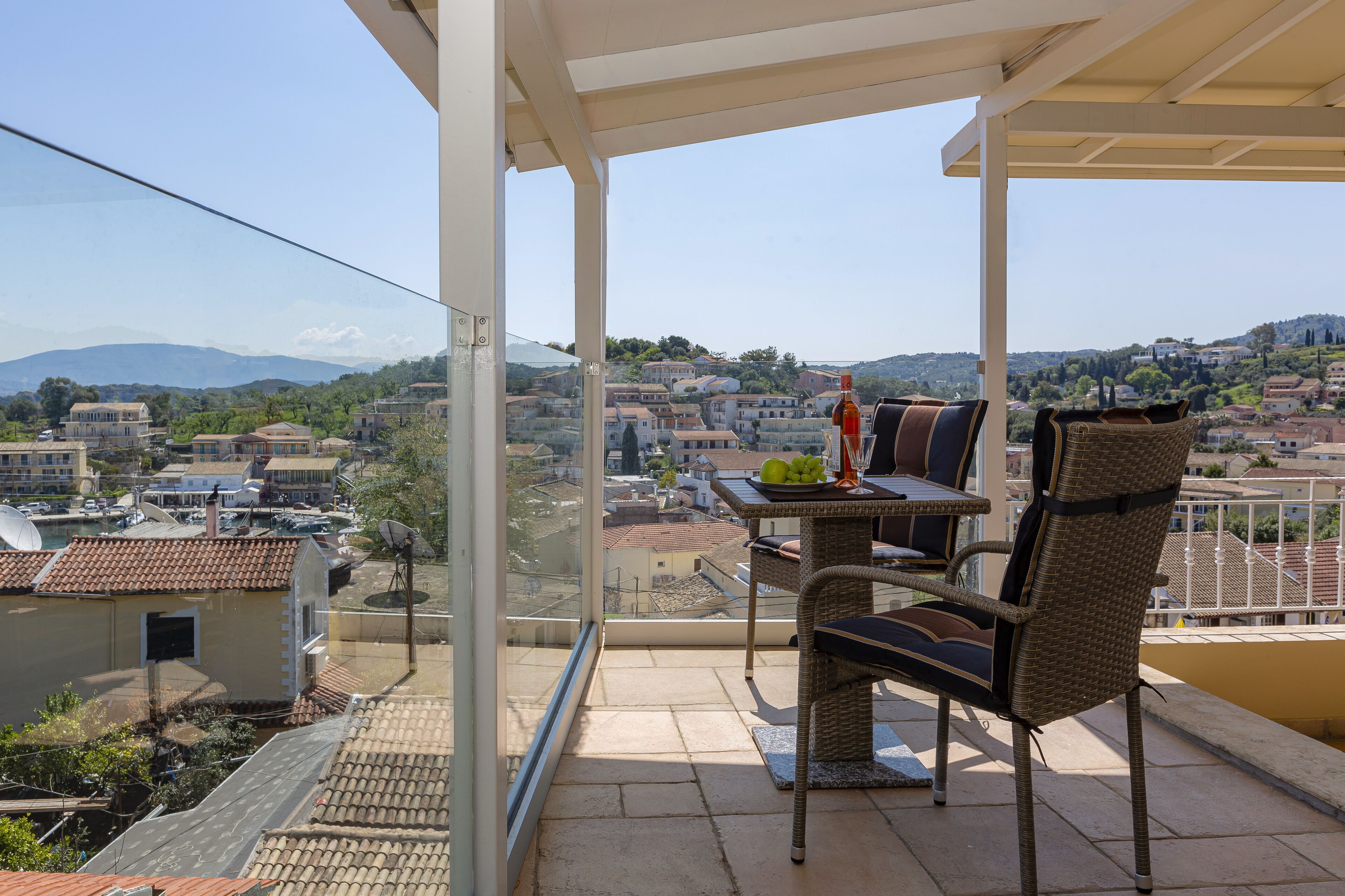 Een balkon met rieten meubels, een tafel met een fruitschaal, en een uitzicht op een stad met huizen en bergen op de achtergrond.