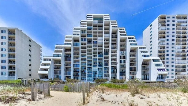Un edificio moderno de gran altura con múltiples balcones y ventanas amplias, ubicado cerca de una zona de playa arenosa.