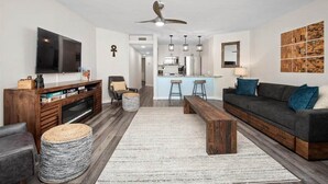 Una sala de estar moderna con sofá, televisor, mueble de entretenimiento de madera y una cocina al fondo.