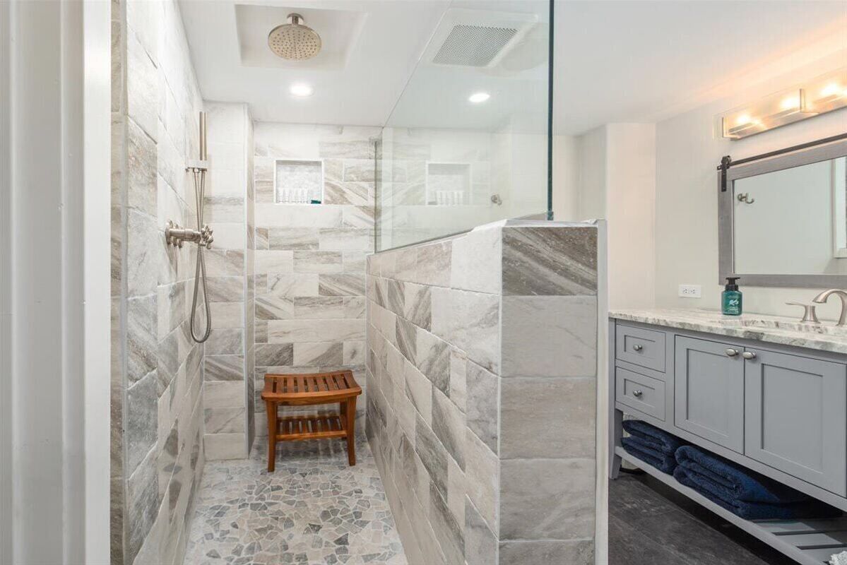 Une salle de bain moderne dotée d’une douche à parois de verre, de carreaux de marbre, d’un banc en bois et d’un meuble-lavabo avec un miroir et un porte-serviettes.