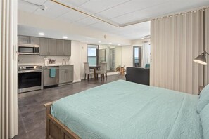A modern bedroom with a large bed, a kitchenette with a microwave and stove, a dining area with a table and chairs, and a view of the sea through a sliding door.