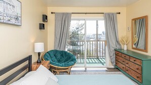 A bedroom with a bed, a chair, a dresser, a lamp, and a large window with curtains.