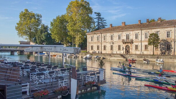 A riverside café with outdoor seating overlooking the water, a historic stone building, and rowboats.