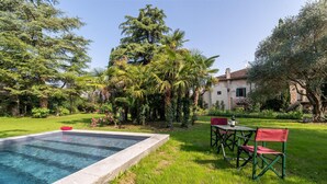 A swimming pool with a table and chairs, surrounded by lush greenery and a building in the background.