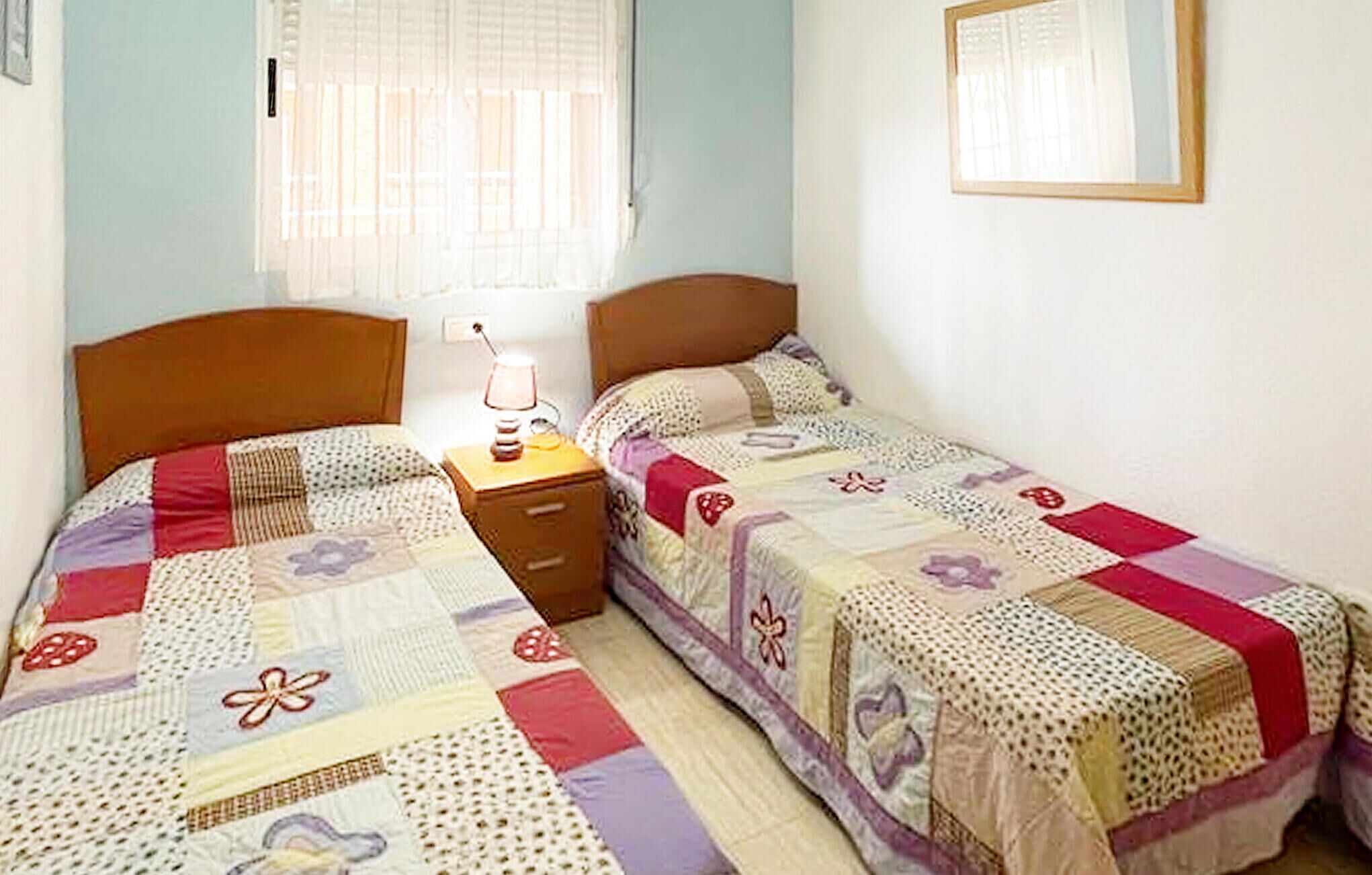 A bedroom with two single beds, a nightstand with a lamp, and a window with blinds.