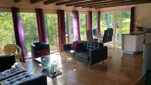 A spacious living room with a leather sofa, a glass coffee table, and a dining area with a table and chairs. Large windows with curtains provide a view of the outdoors. - eco-gîte with SPA in the heart of nature, horse riding, Limousin (DOMPIERRE-EGLISES)