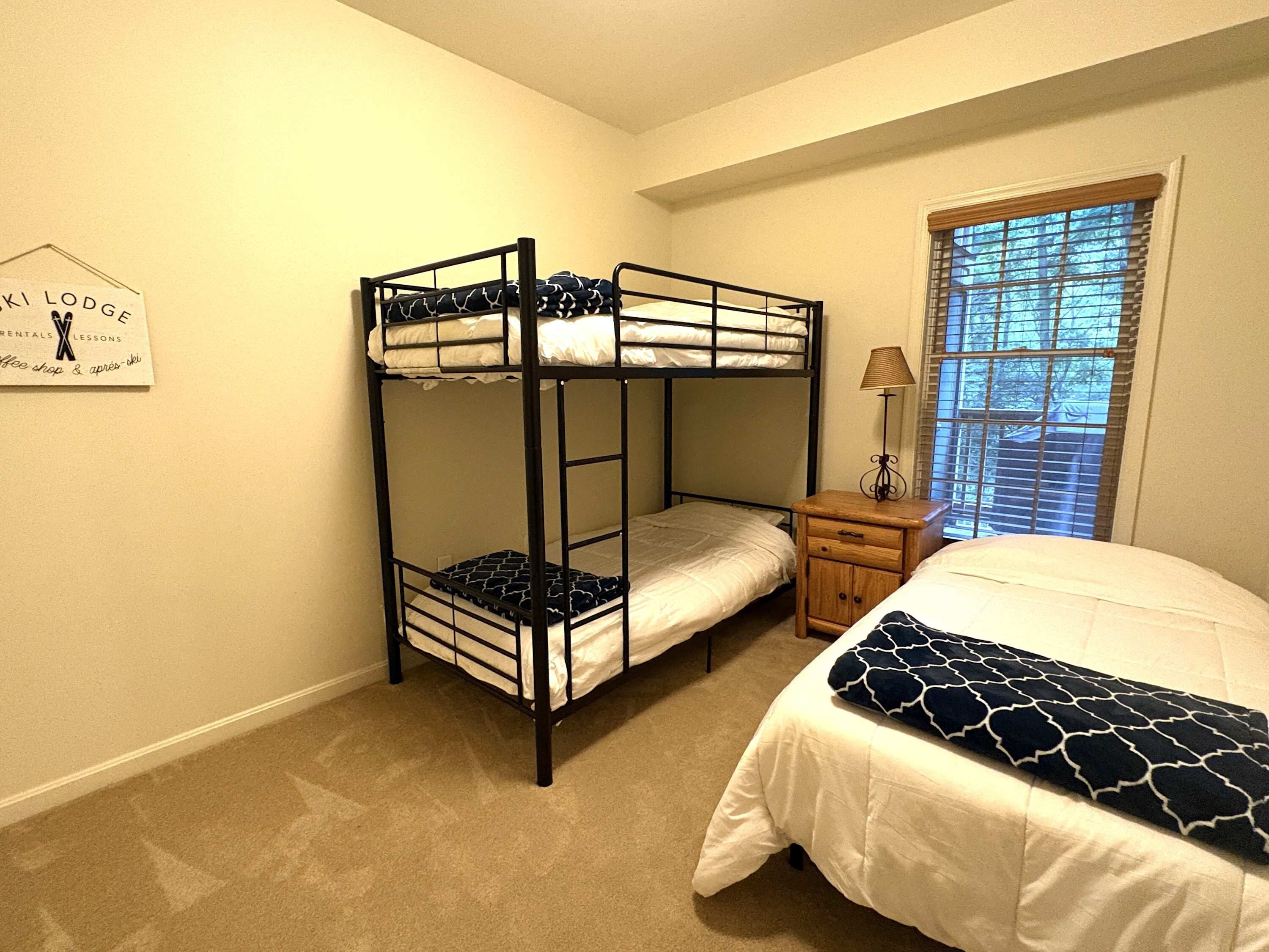 A bunk bed room with a single bed, a nightstand with a lamp, and a window with blinds.