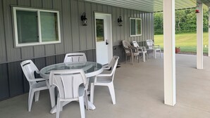 A covered patio with a round glass table and white plastic chairs, a door, and windows with shutters. - UpNorth Getaway with access to ATV UTV Snowmobile Trail (Birchwood)