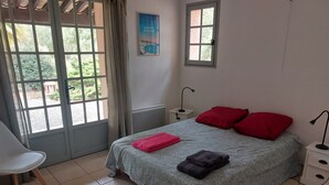 A bedroom with a bed, two red pillows, a nightstand with a lamp, a chair, and a window with a view of greenery. - Villa de prestige au cœur d’un domaine privé
Piscine privée & sérénité absolue (Le Muy)