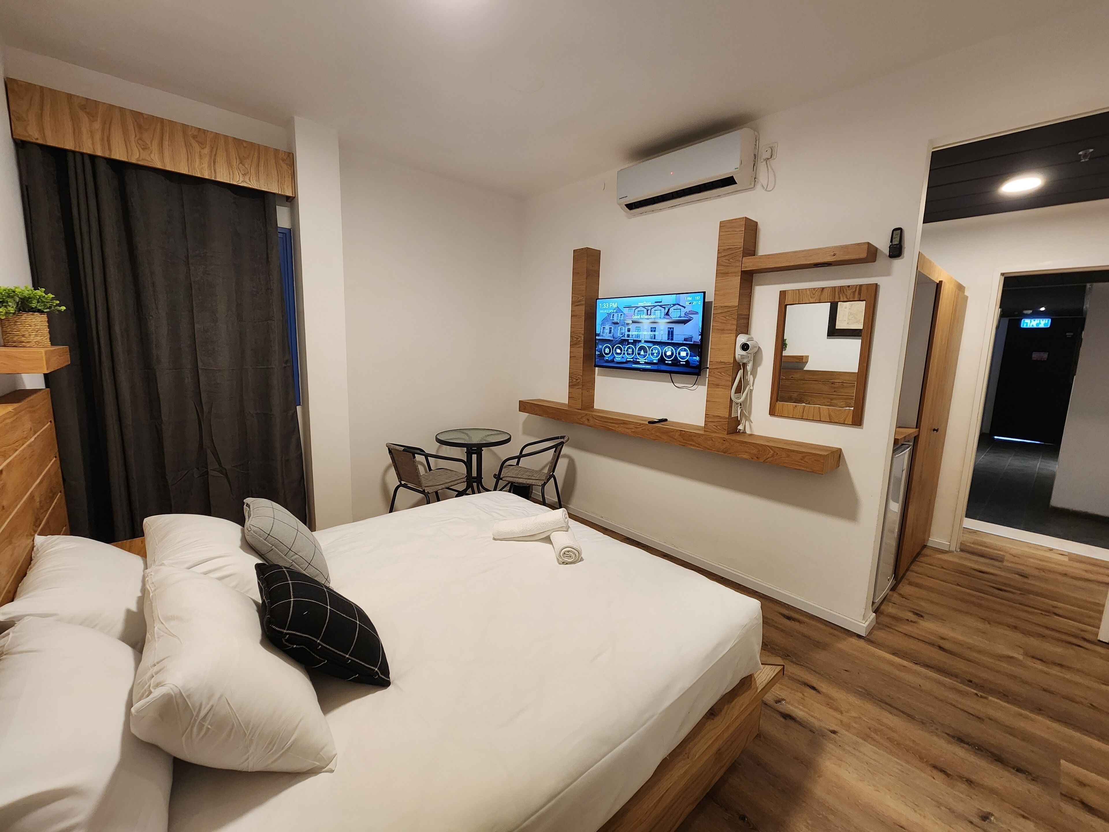 A bedroom with a bed, a small table with chairs, a mounted TV, an air conditioning unit, and a wooden floor.
