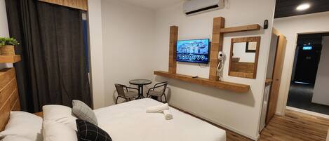 A bedroom with a bed, a small table with chairs, a mounted TV, an air conditioning unit, and a wooden floor.