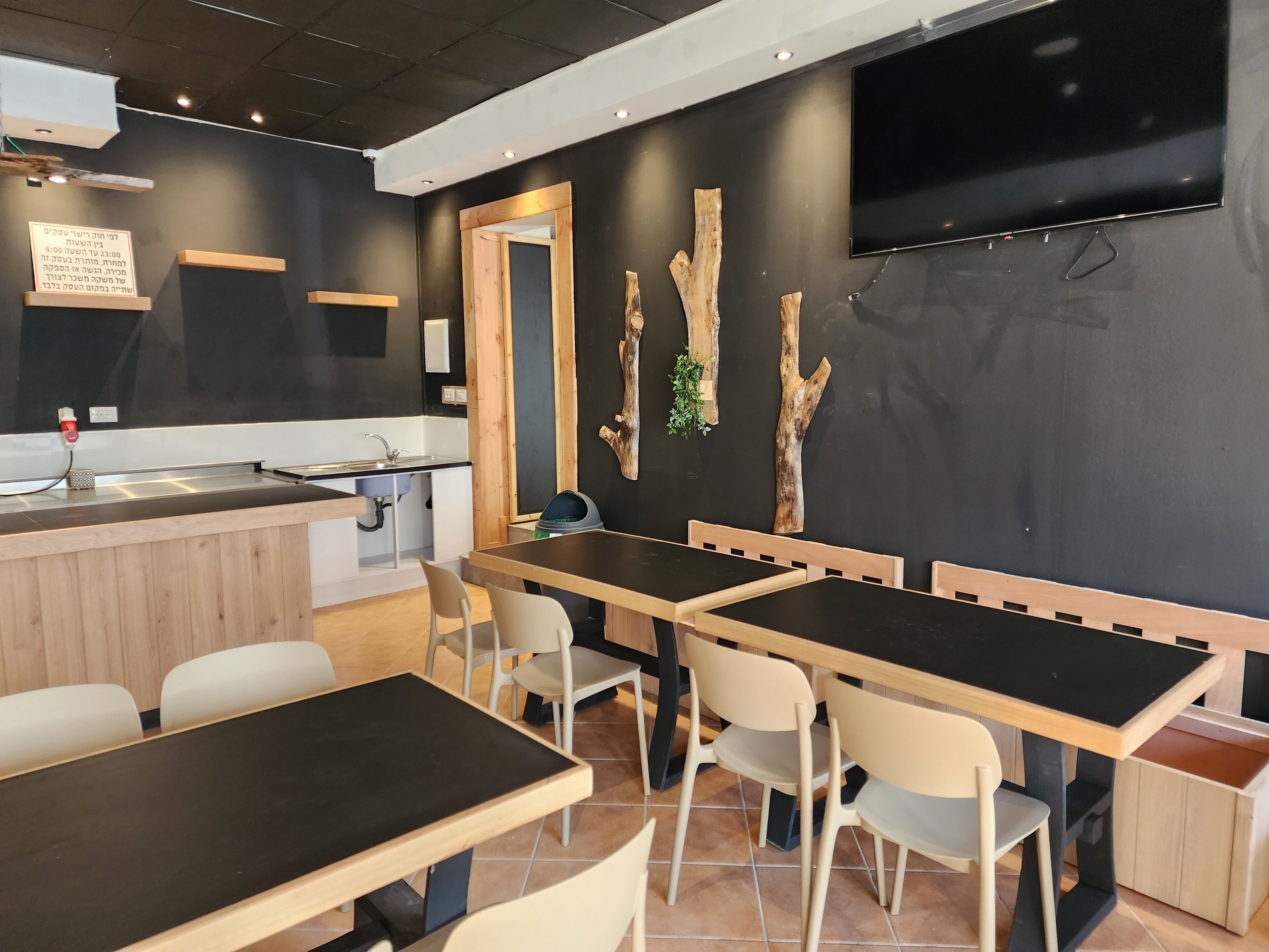 A modern restaurant interior with wooden tables and chairs, a black countertop with a sink, and a mounted television on the wall.